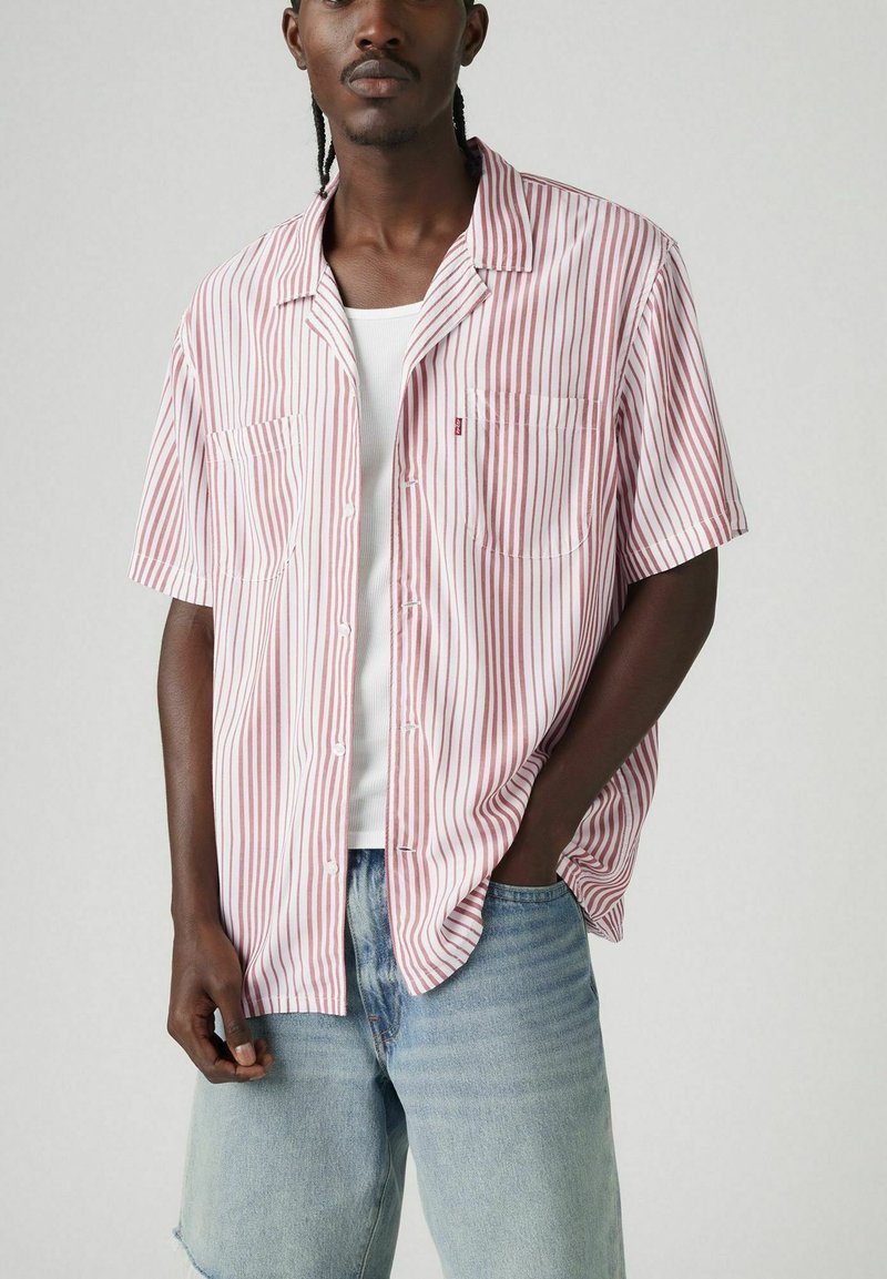 Camisa de manga corta a rayas en blanco y rojo, hecha de tela ligera, con cierre de botones en la parte delantera y dos bolsillos en el pecho. Llevada sobre una camiseta blanca básica.
