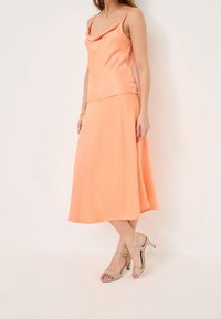 Femme portant un top sans manches en satin pêche avec un col bénitier et une jupe midi assortie, associée à des sandales à talons hauts beige.