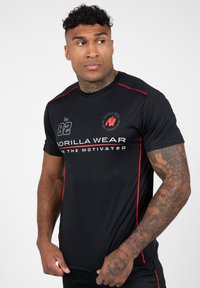 Zwarte sport t-shirt met rode accenten, met een ronde hals, korte mouwen en bedrukte logo's en nummers op de voorkant en mouw.