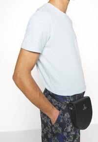 Chemise bleu clair à manches courtes associée à un pantalon sombre à motifs et un sac bandoulière noir incurvé avec des éléments en métal.