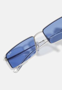 Rektangulära solglasögon med blå linser, silvermetallram, klara näskuddar och en strömlinjeformad design. "Ray-Ban"-logotyp på linsen.