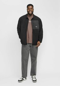 Sort fleecetrøje med lynlåslomme på brystet, brun t-shirt under, grå bukser og sorte og hvide sneakers. Står mod en hvid baggrund.