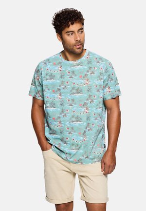 Hombre con camisa de manga corta estampada tropical turquesa y pantalones cortos beige remangados, posando con una mano en el bolsillo, mirando hacia la derecha.