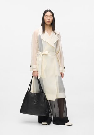 Lagana, prozirna baloner jakna u krem boji sa sivim detaljima, uparena s crnom kožnom torbom s istaknutim logotipom i širokim crnim hlačama.
