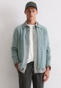 Jasnoniebieska polarowa kurtka z kołnierzem, z guzikami na froncie i kieszenią na piersi, noszona na koszuli w kratę. Szare spodnie i zielona czapka.