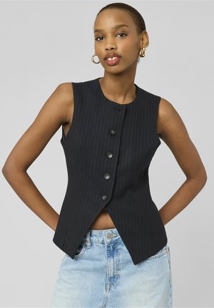 Femme portant un gilet noir rayé sans manches boutonné, associé à un jean en denim bleu clair, posant avec les mains derrière le dos.