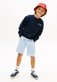 Μπλε φούτερ με το λογότυπο "TOMMY", ανοιχτό μπλε σορτς, κόκκινο καπέλο τύπου bucket, μπλε παπούτσια με λευκά κορδόνια και λευκές κάλτσες με σχέδια. Casual εμφάνιση.