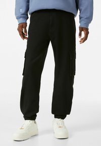 Pantaloni cargo neri con tasche laterali, polsini elasticizzati e vestibilità comoda, abbinati a sneakers bianche e una felpa blu.