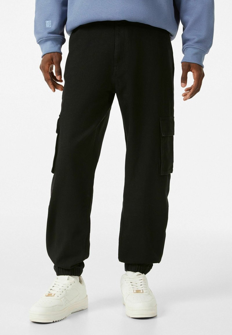 Pantaloni cargo neri con tasche laterali, polsini elasticizzati e vestibilità comoda, abbinati a sneakers bianche e una felpa blu.