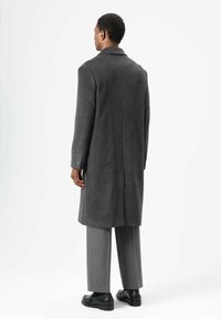 Grauer Wollmantel mit maßgeschneidertem Design, ausgestattet mit einem Reverskragen und Manschetten mit einem Knopf, kombiniert mit grauer Hose und schwarzen Schuhen.