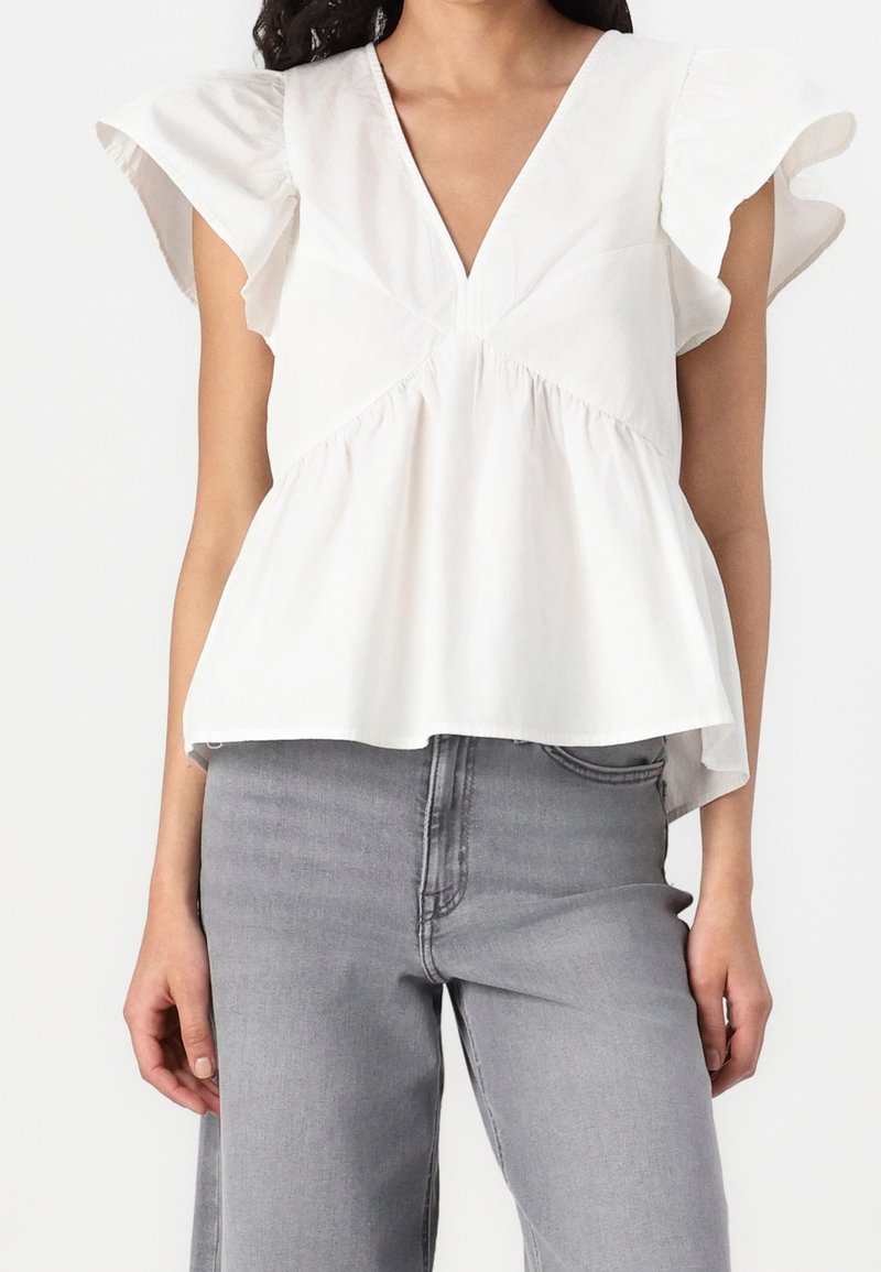 Femme portant un chemisier blanc à manches volantées avec un décolleté en V profond, associé à un jean gris taille haute sur un fond uni.