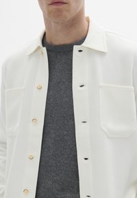 Hvid skjortejakke med krave, udstyret med lysebrune knapper, en brystlomme, over en mørkegrå sweater. Glat stofstruktur.
