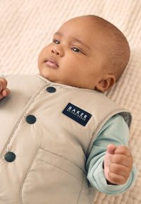 Veste matelassée beige avec des boutons noirs et un patch logo. En dessous, chemise à manches longues bleu clair. La main du bébé est serrée. Fond à texture douce.