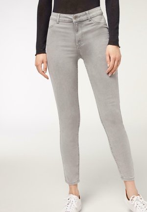 Person trägt hellgraue, schlanke Jeans, schwarzen langärmligen Oberteil und weiße Sneaker, steht vor neutralem Hintergrund.