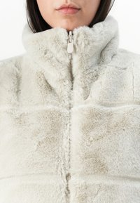 Faux fur jas in lichtbeige met een hoge kraag en een ritssluiting aan de voorkant. De textuur van de stof heeft een zachte, pluche oppervlakte met een doorgestikt ontwerp.