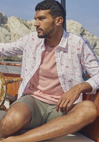 Zittende man in een wit shirt met flamingopatroon over een roze crewneck t-shirt, groene shorts, op een boot met een rotsachtige achtergrond.