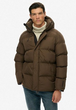 Mand iført en brun polstret jakke med hætte, beige rullekravesweater og blå jeans, stående mod en ensfarvet hvid baggrund.