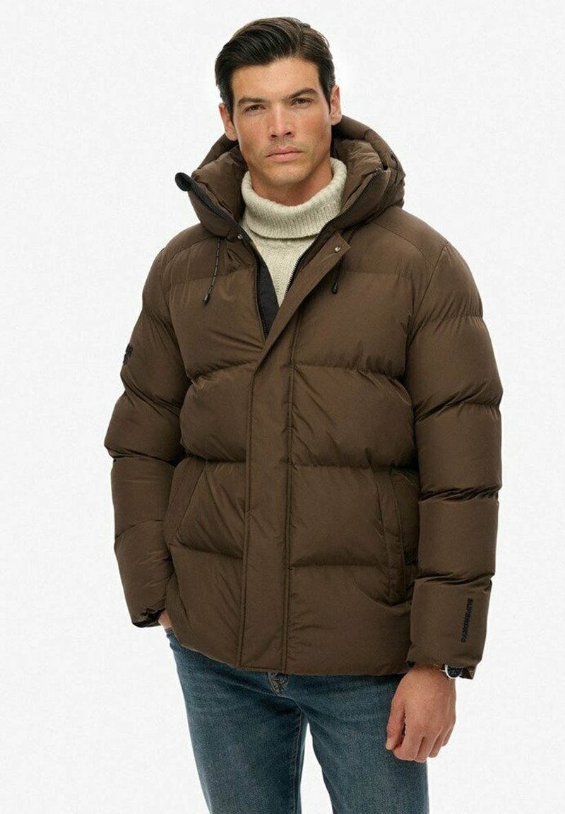 Mand iført en brun polstret jakke med hætte, beige rullekravesweater og blå jeans, stående mod en ensfarvet hvid baggrund.