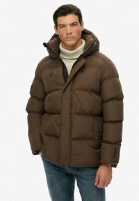 Brun oppustet jakke med hætte, der har en lynlåslukning foran, store quiltede sektioner og sidelommer. Båret over en lys sweater.