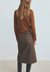 Pull en maille marron à manches longues associé à une jupe plissée marron à carreaux, longueur genou. Bottes noires montant jusqu'aux genoux complètent la tenue.