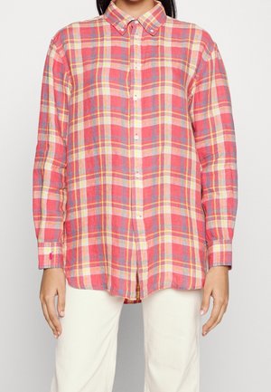 Femme portant une chemise à manches longues à carreaux rouge, rose, jaune et blanc avec un pantalon de couleur claire.