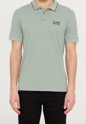 Mężczyzna noszący jasnozieloną koszulkę polo EA7 Emporio Armani z czarną lamówką na kołnierzyku i czarne spodnie, stojący na jednolitym tle.