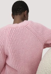 Person von hinten zu sehen, der einen strukturierten rosa Strickpullover mit gerippten Mustern vor einem schlichten hellen Hintergrund trägt.