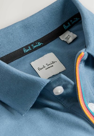 Mėlyna polo marškinė su reljefiniu paviršiumi, baltais mygtukais ir apykakle su daugiaspalviais krašteliais. Etiketėse užrašyta „Paul Smith“ ir „JUNIOR“. 
