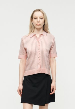 Młoda kobieta w różowym, krótkim sweterku na guziki z cienkimi paskami i czarnej spódnicy, stojąca na jednolitym tle.