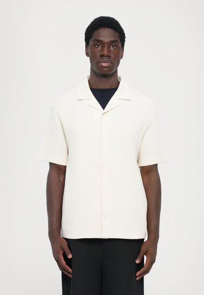 Chemise à manches courtes, col classique en tissu crème clair avec une texture, présentant un devant à boutons et une coupe décontractée, assortie à un pantalon sombre.
