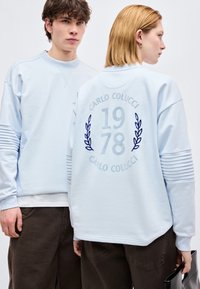 Δύο μοντέλα με ανοιχτό μπλε φούτερ· το ένα κοιτάει μπροστά, το άλλο δείχνει την πλάτη με το κείμενο "Carlo Colucci 1978" και σχέδια δάφνης.