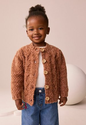 Brun strikket cardigan med et tekstureret mønster, træknapper, båret over en hvid top, parret med denimjeans med blomsterbroderi.