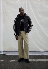 Črna jakna z zadrgo spredaj in belimi črtami na straneh, črna majica, bež hlače širokih nogavic, črni čevlji, stoji pred svetlim ozadjem.