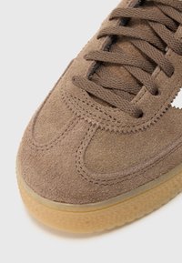 Καφέ σουέντ αθλητικό παπούτσι με στρογγυλερή μύτη, υφή στην επι�φάνεια, επίπεδα κορδόνια και σόλα από καουτσούκ. Απλές ραφές και διάτρητες λεπτομέρειες διακοσμούν το σχέδιο.