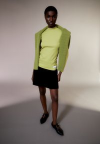 Groene gebreide trui over een nauwsluitend gele top, gecombineerd met een zwarte fluwelen minirok en puntige zwarte schoenen met gespsluiting.