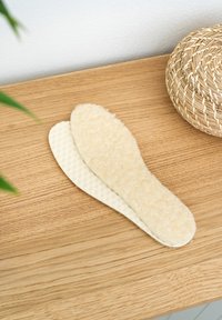 Cream-colored insoles with a textured surface and a soft, fuzzy layer on top, placed on a wooden surface, near a woven basket.