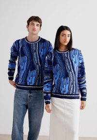 Deux modèles portent des pulls en tricot à motifs bleus avec des manches longues, présentant des designs contrastés en bleu foncé et en bleu clair. L'un porte un jean, l'autre une jupe blanche.
