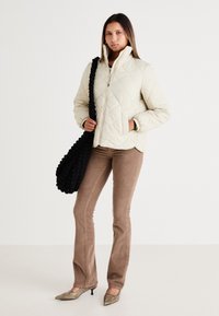 Lys beige quiltet jakke med høj krave, parret med brune kordfløjl bukser og metalliske spidse hæle. Sort struktureret skuldertaske.