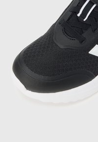 Close-up af tåen på en sort atletiksneakers med mesh-overdel, sorte snørebånd og hvid polstret sål på en lys baggrund.