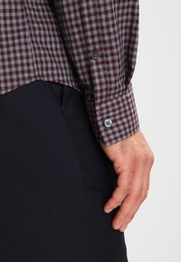 Camicia a quadri bordeaux e grigia con polsini abbottonati, abbinata a pantaloni scuri. Presenta una trama liscia e colori a contrasto.