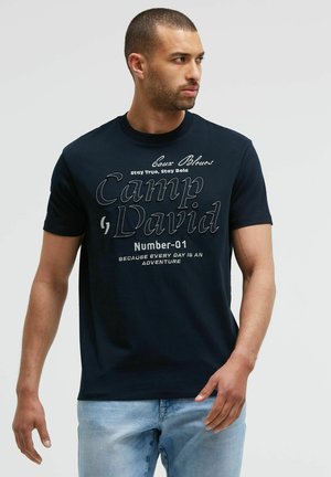 Mann mit kurzen Haaren, der ein schwarzes "Camp David" T-Shirt und hellblaue Jeans trägt und nach rechts schaut vor einem schlichten Hintergrund.