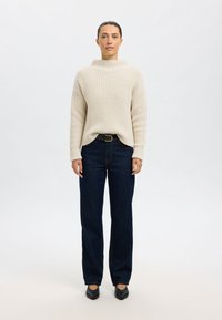 Pull beige côtelé à col montant, associé à un jean large bleu foncé et des ballerines noires. Tenue simple et décontractée avec des accents minimes.