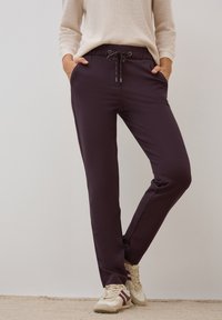 Pantalon de jogging violet foncé avec taille élastique et cordon de serrage, présentant des poches latérales et une coupe fuselée. Porté avec des baskets beiges.