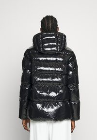 Osoba s copy v vlasech, oblečená v lesklé černé péřové bundě s kapucí, otočená zády, na jednobarevném bílém pozadí.