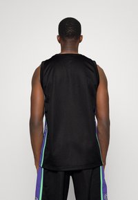 Muž viděný zezadu, oblečený v černém bezrukávovém basketbalovém dresu s fialovými a zelenými bočními panely a odpovídajících šortkách.