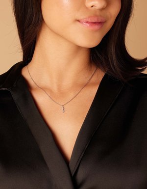 Collier en argent avec une chaîne délicate, mettant en avant de petites pierres précieuses scintillantes disposées en ligne verticale. Porté sur un haut en satin noir.