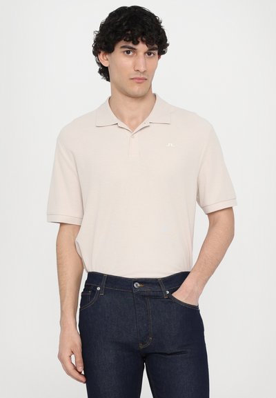 Cămașă polo bej cu mâneci scurte și guler clasic, având un logo subtil pe piept; asortată cu blugi din denim de culoare albastru închis.