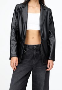Femme portant un blazer noir en similicuir, un crop top blanc et un jean à jambes larges gris foncé taille haute, debout devant un fond blanc.