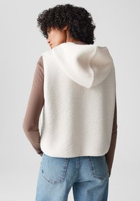 Weißer Fleece-Weste mit Kapuze, die eine strukturierte Oberfläche, einen abgerundeten Saum hat und über einem braunen Langarmshirt getragen wird, kombiniert mit blauen Jeans.
