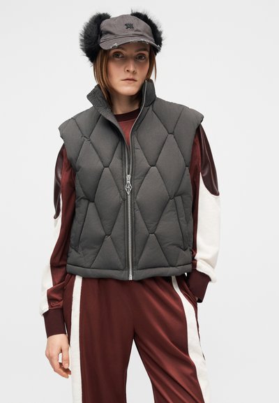 Szürke párnázott mellény gyémánt alakú steppeléssel, elöl cipzárral és magas gallérral, bordó hosszú ujjú felső és hozzá illő, fehér csíkos nadrág fölött viselve.